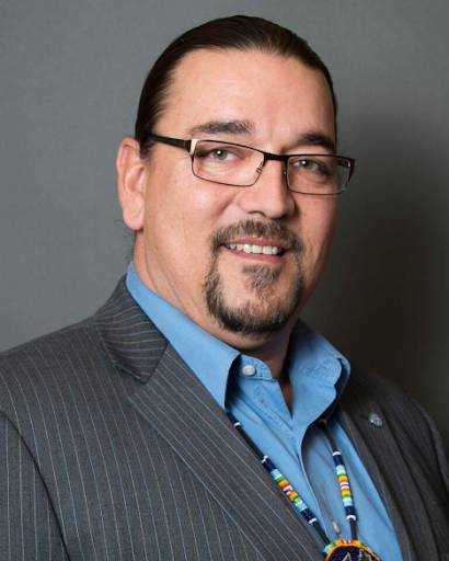 The image shows a man with long, dark hair pulled back, wearing glasses and a goatee. He is dressed in a pinstripe suit jacket over a blue button-down shirt, accessorized with a beaded necklace in traditional Native American style. He has a friendly expression and is looking at the camera.